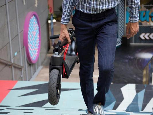 Man in business casual attire carrying a lightweight folded electric scooter up a set of urban stairs