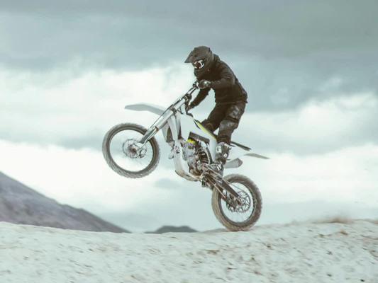 Rider performing a wheelie jump on a white electric dirt bike against a cloudy sky on a sandy hill