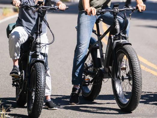 Close-up of two people riding black fat tire electric bikes on a paved road, focusing on the front suspension forks and tires