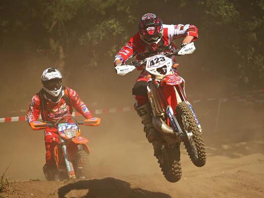 Two motocross riders racing on a dirt track, with the lead rider performing a jump on a red dirt bike