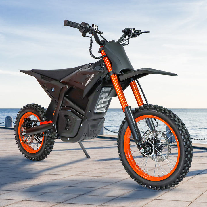 A front-quarter studio shot of a black electric dirt bike featuring vibrant orange front forks and wheel rims, parked on a wooden dock with a calm ocean background.