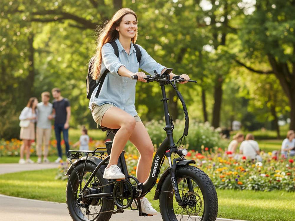 Young woman smiling while riding a black Cemoto folding electric bike with fat tires on a paved path in a sunny park
