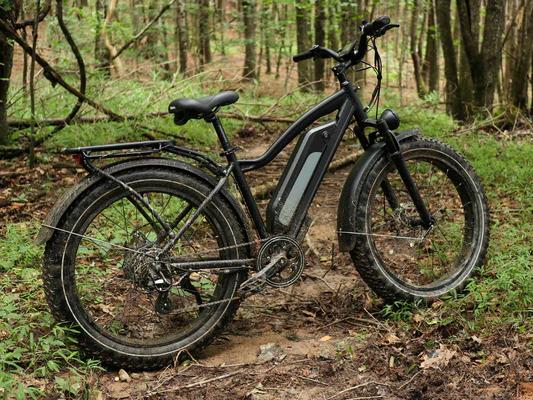 Black Cemoto electric hunting bike with fat tires and rear rack parked on a muddy trail in a dense forest