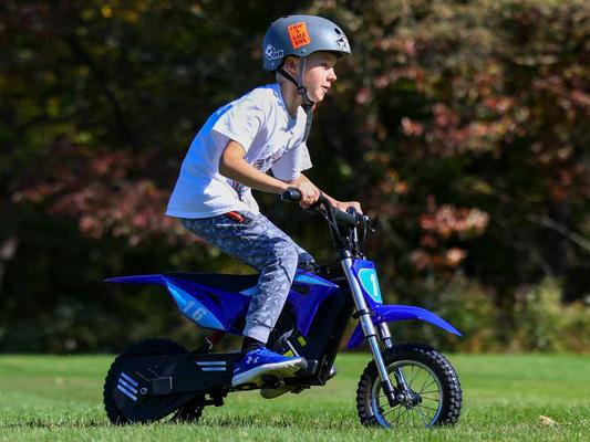 Young boy wearing a helmet riding a blue mini electric dirt bike on a grassy field