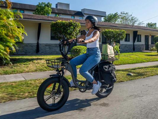 Woman riding a black cargo electric bike with front and rear baskets filled with groceries on a residential street