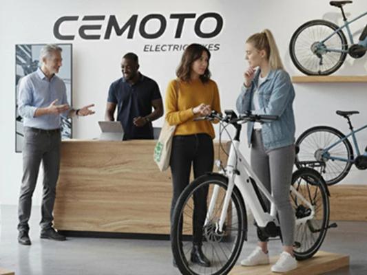 Salesperson showing a white Cemoto electric bike to a customer in a modern bike shop with the brand logo on the wall